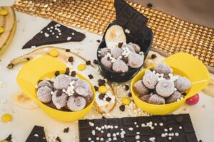 Mini Melts-ის შოკოლადის ნაყინის ბურთულები ყვითელ და შავ ჭურჭელში, გარშემო შოკოლადის ფილებითა და ბანანის ნაჭრებით.