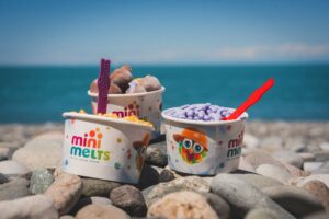 Mini Melts-ის სხვადასხვა ფერის ნაყინის ჭიქები ფერადი კოვზებით ზღვის სანაპიროზე, კენჭებზე.